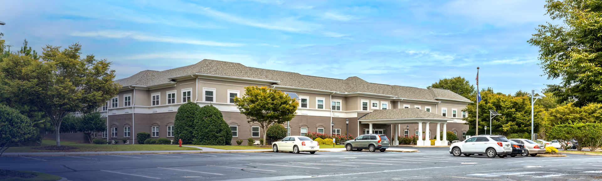 Front exterior of a two-story senior living facility with a covered entrance, parked cars, and landscaped trees.