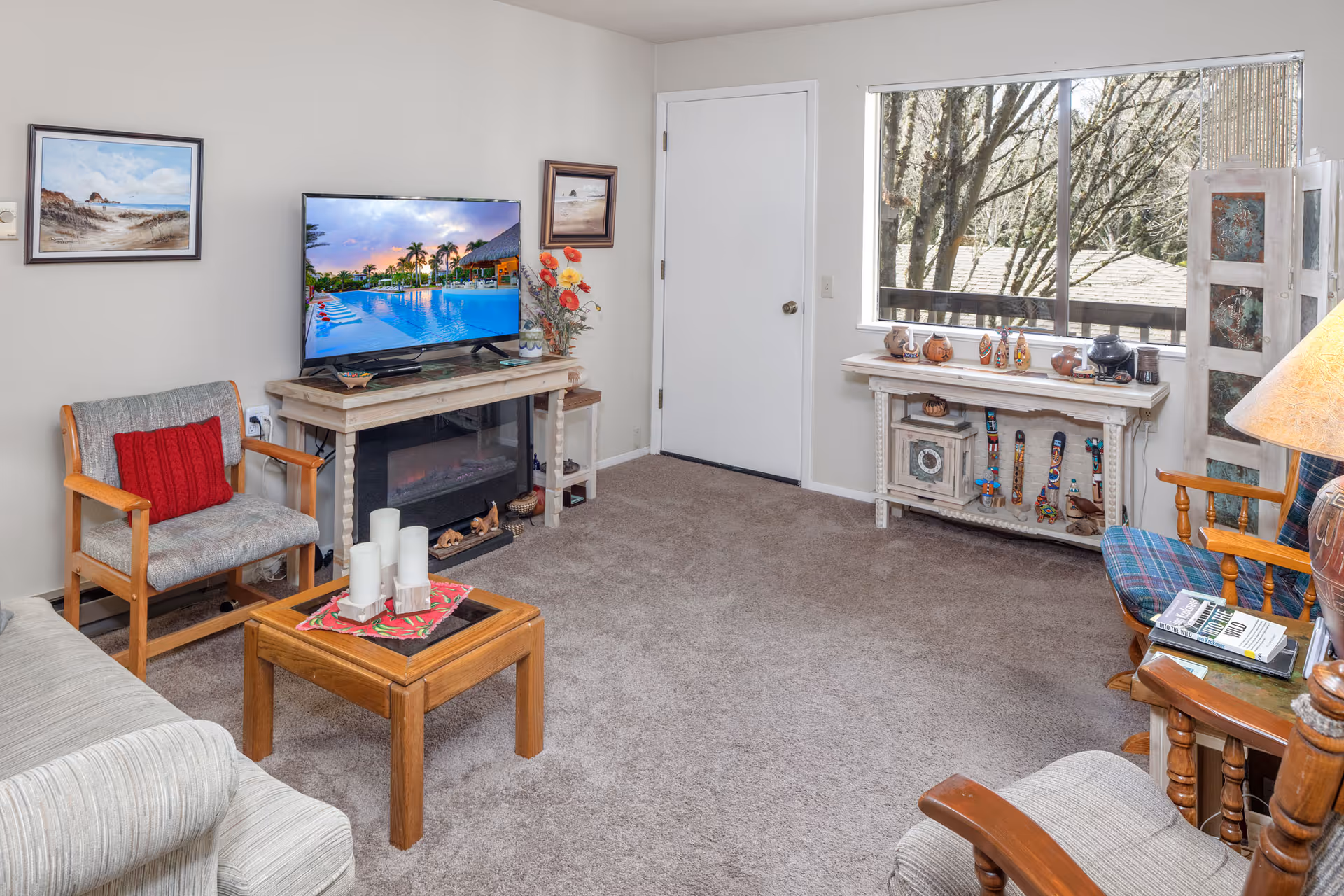 A cozy living room with beige carpet and light-colored walls. The room features a TV on a wooden stand above an electric fireplace, a wooden armchair with a red cushion, a small wooden coffee table with white candles, and a window showing trees outside. There are decorative items on a white console table beneath the window, framed paintings on the walls, and a floor lamp beside a plaid cushioned chair.
