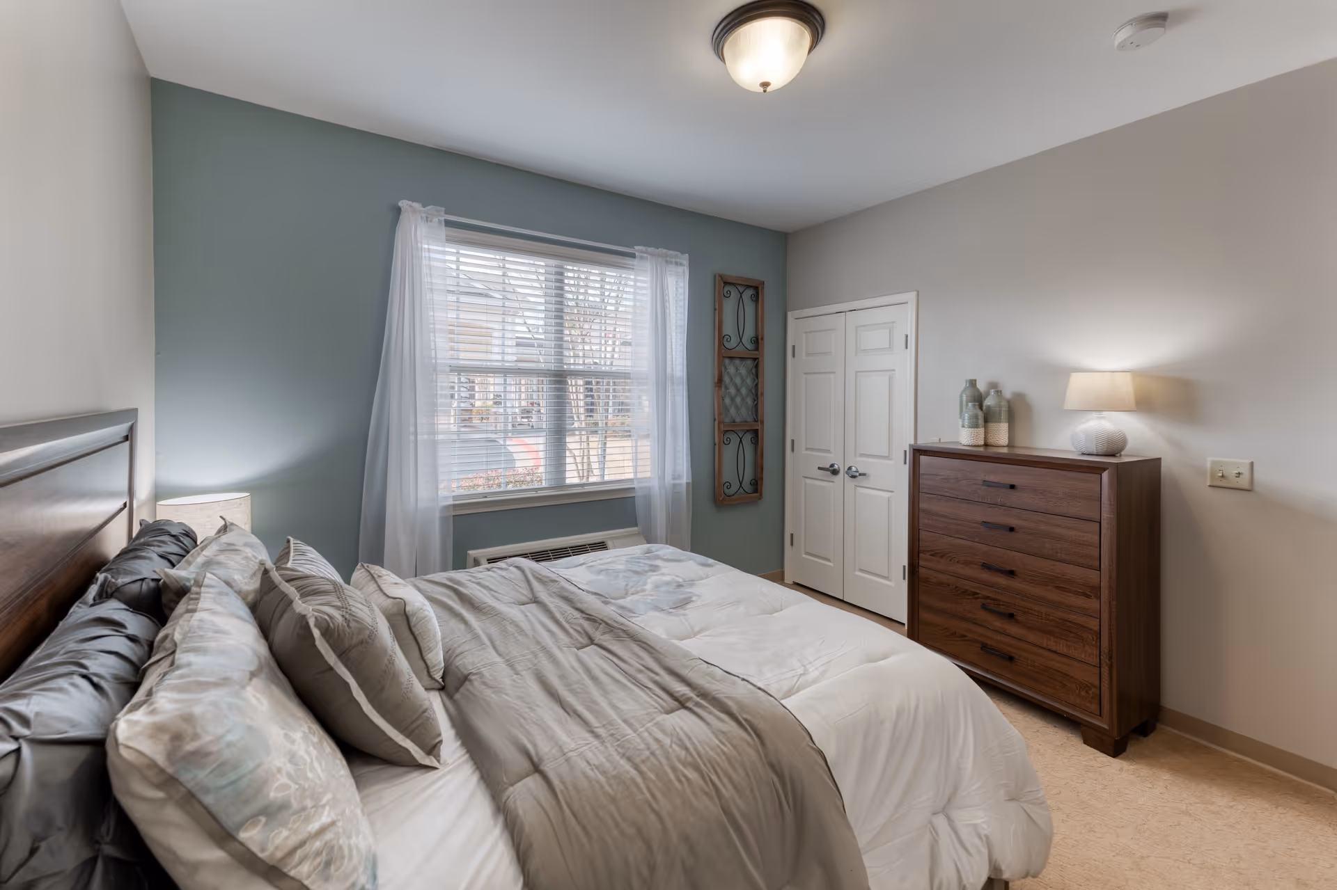 A neatly made bed with multiple pillows in a bedroom featuring a large window with sheer white curtains, a wooden dresser with decorative vases and a lamp, and a double-door closet. The walls are painted in light gray and muted green tones.
