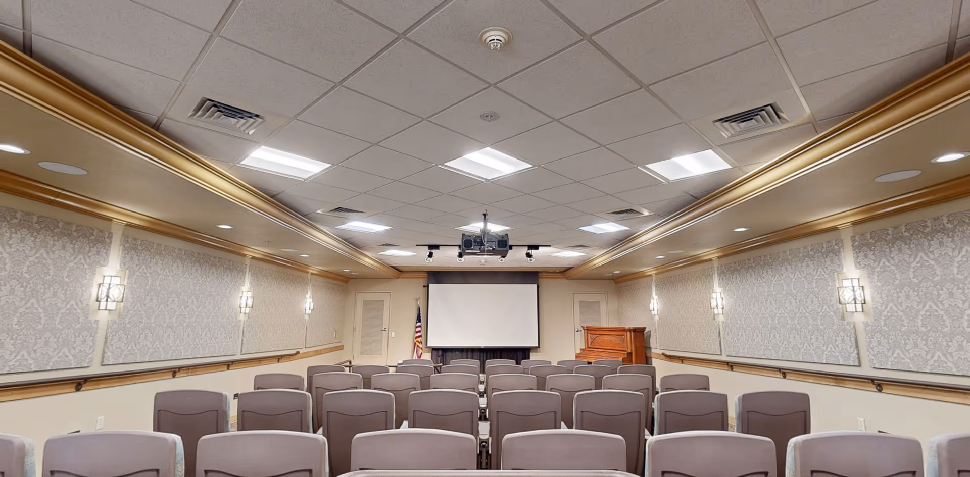 A conference or presentation room with rows of gray chairs facing a large white projection screen. The room has patterned wallpaper, wall sconces with lights, a wooden podium, and an American flag near the screen. The ceiling has recessed lighting and a mounted projector.