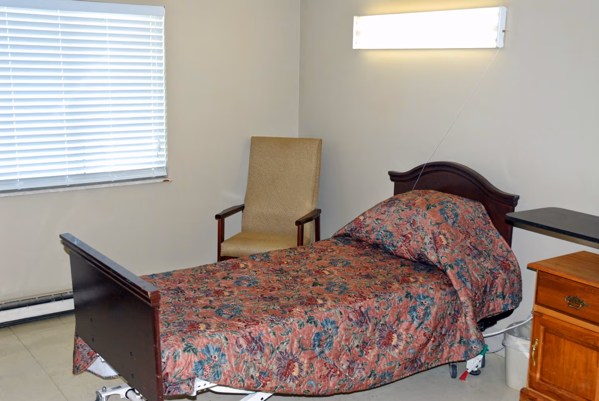 A small room with a single hospital-style bed covered with a floral patterned bedspread. Next to the bed is a wooden nightstand with a drawer and a beige upholstered chair is placed against the wall near a window with closed blinds. A bright rectangular light fixture is mounted on the wall above the bed.
