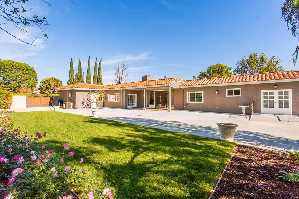 Outdoor view of a single-story building with a tiled roof, surrounded by a well-maintained lawn and flower beds under a clear blue sky.