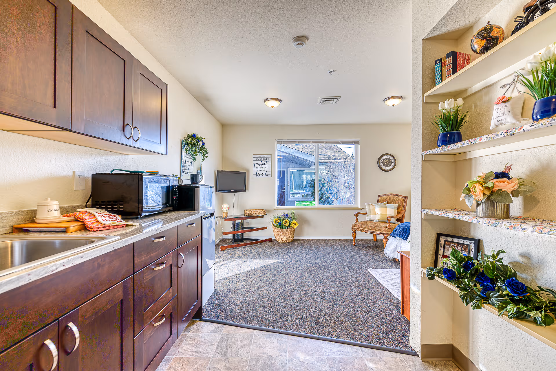 A bright and cozy interior space featuring a kitchenette with dark wooden cabinets, a microwave, and a small refrigerator. The room extends into a carpeted living area with a window, a decorative chair with pillows, a small TV on a stand, and various decorative plants and flowers on shelves and in baskets. The walls are light-colored, and the space is well-lit with ceiling lights and natural light from the window.