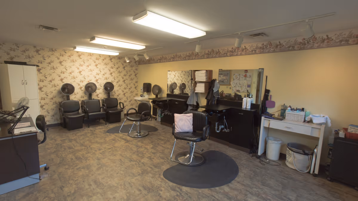 Interior of a hair salon room with three black hair drying chairs against a floral wallpapered wall, two black salon chairs with sinks and mirrors, a white cabinet, and a small white table with various hair care products and towels.