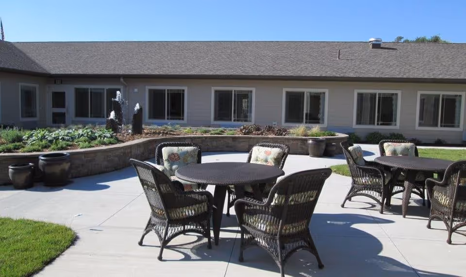 Outdoor patio area with round tables and wicker chairs with cushions, situated on a concrete surface. In the background, there is a low stone wall with plants and a small water fountain, and a single-story building with multiple windows under a clear blue sky.
