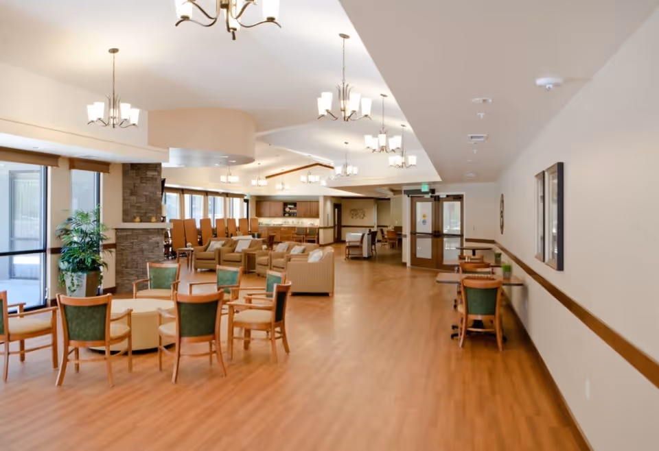 A spacious senior living facility common area with wooden flooring, multiple seating arrangements including green cushioned chairs around tables, beige sofas, and armchairs. The room features large windows allowing natural light, a stone fireplace, and several ceiling light fixtures. There is a doorway at the far end and some framed artwork on the walls.