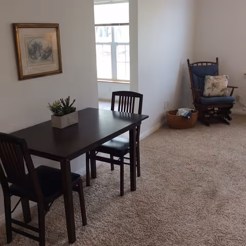 Carpeted living area with a small dark wood dining table and chairs, a rocking chair by the wall, framed art, and a window-lit alcove.