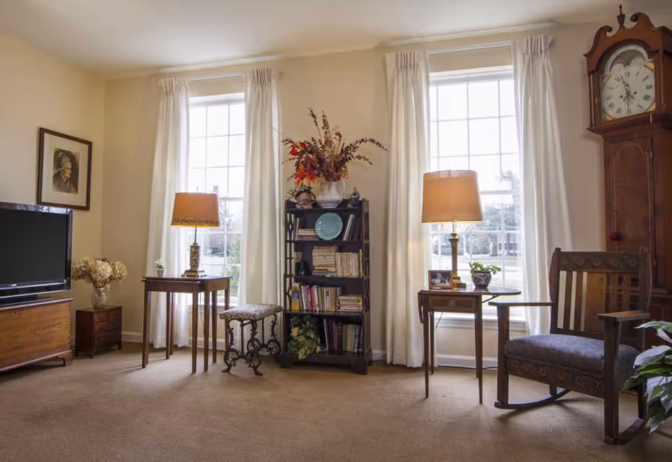 A cozy living room with two large windows covered by white curtains, a wooden rocking chair, a tall grandfather clock, a bookshelf filled with books and decorative items, two side tables each with a lamp, a flat-screen TV on a wooden stand, and a small stool with a floral cushion.