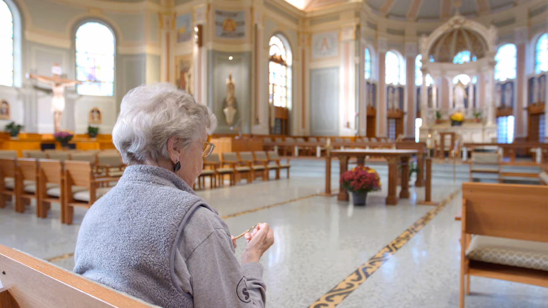 An elderly woman with gray hair and glasses sitting on a wooden pew inside a large, ornate chapel or church, holding a rosary. The chapel features stained glass windows, religious statues, and wooden benches arranged in rows.