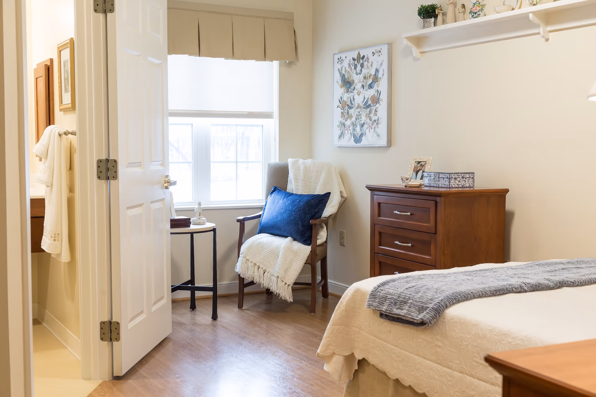 A cozy bedroom in a senior living facility featuring a bed with a white quilt and gray throw, a wooden dresser with decorative items, a wooden chair with a white blanket and blue pillow, and a small round side table near a window with beige curtains. The door to an adjacent bathroom is open, showing a towel hanging inside.