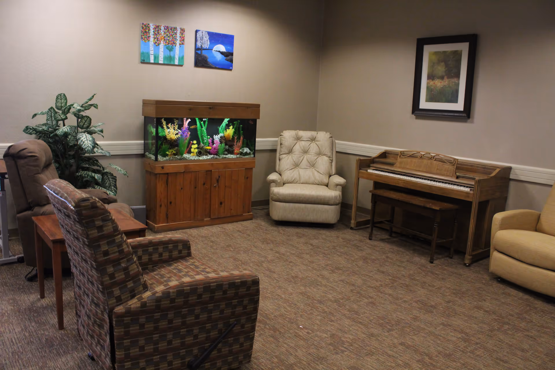A cozy sitting area with four armchairs arranged around a wooden fish tank filled with colorful artificial plants. There is a wooden piano with a bench against the wall, a potted plant in the corner, and three framed artworks hanging on the walls. The room has beige walls and carpeted flooring.
