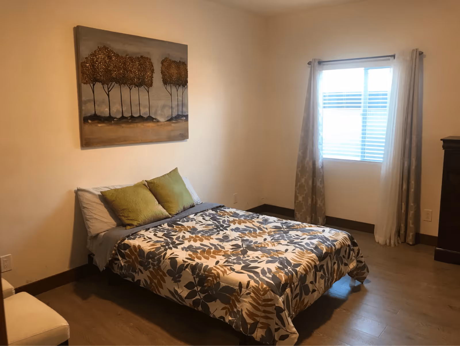 A bedroom with a bed covered in a floral patterned comforter with blue, brown, and beige leaves. Two green pillows rest against white pillows. Above the bed hangs a painting of trees with brown foliage. To the right, there is a window with blinds and beige curtains. The floor is wooden, and a dark wooden dresser is partially visible on the right side.