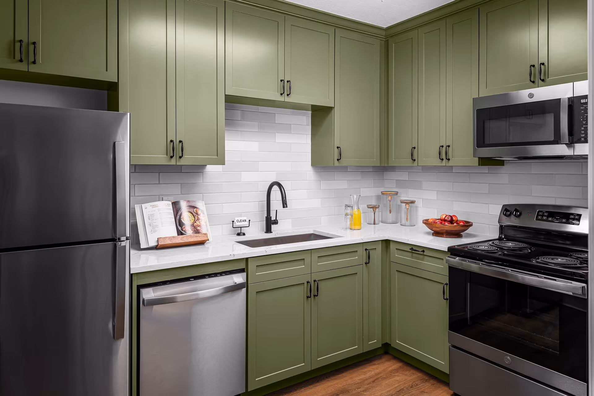Modern kitchen with olive green cabinets, stainless steel refrigerator, dishwasher, microwave, and stove. White subway tile backsplash and white countertop with a black faucet. On the counter are a cookbook on a stand, a small sign that says CLEAN, glass containers, a pitcher with orange juice, and a bowl of red apples.