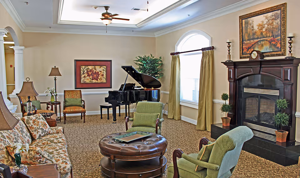 A cozy living room with floral patterned sofa, green upholstered chairs, a round leather ottoman, a grand piano, a fireplace with a wooden mantle, and framed artwork on the walls. The room has beige walls, carpeted floor, and a large window with curtains allowing natural light.