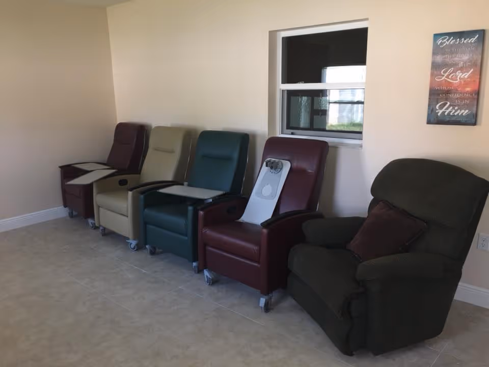 A row of five recliner chairs in different colors lined up against a beige wall in a room with tiled floor. Four of the chairs have small attached trays, and one chair has a brown cushion. There is a small window above the chairs and a framed inspirational sign on the wall.
