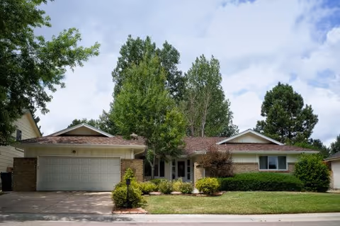 Single-story residential building with a two-car garage, surrounded by trees and shrubs under a partly cloudy sky.