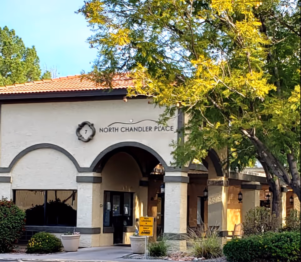 Exterior view of North Chandler Place building entrance with arched doorways, beige walls with gray trim, surrounded by green trees and shrubs under a clear blue sky.