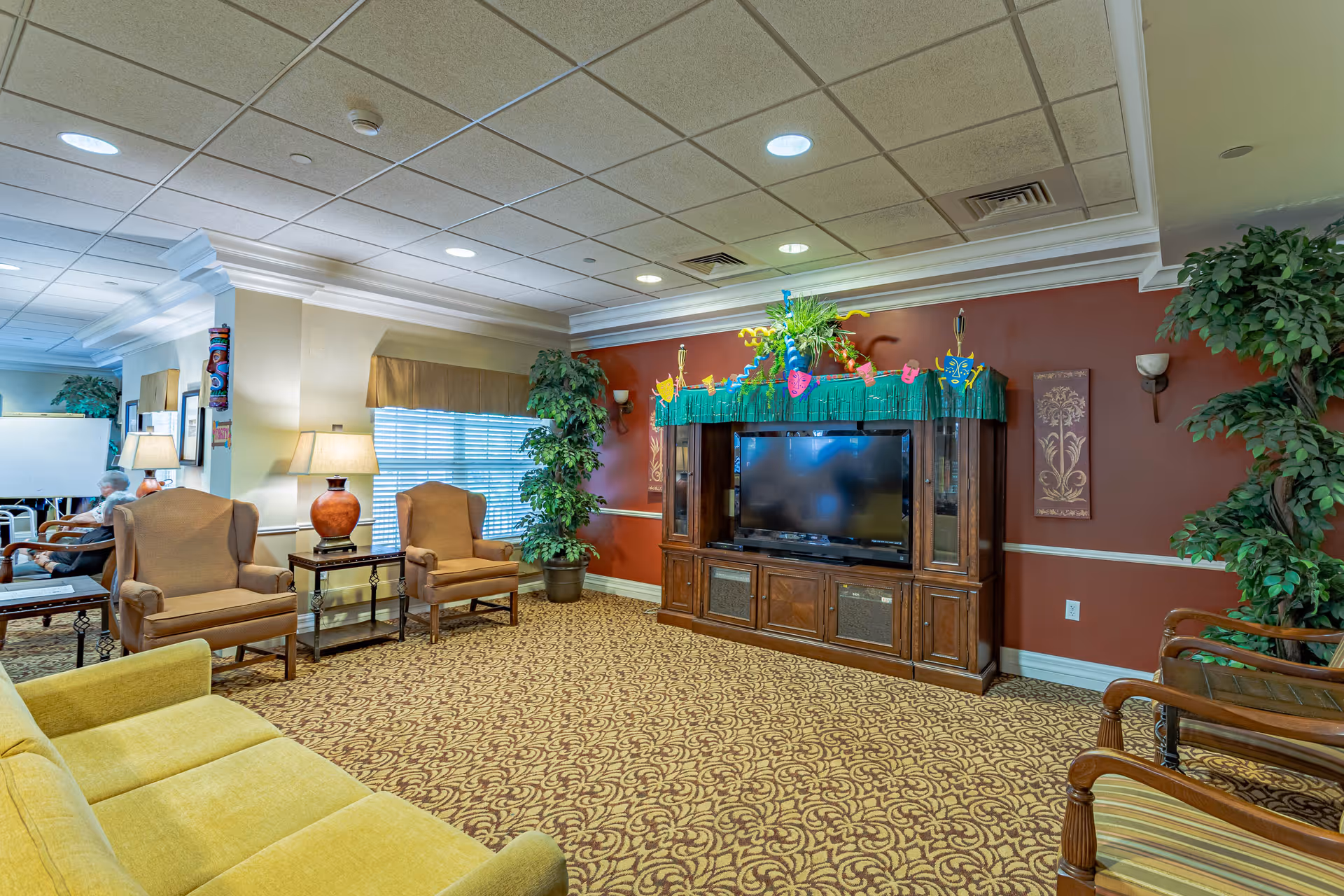 A cozy senior living facility common area with a yellow sofa, two beige armchairs, a wooden entertainment center with a flat-screen TV, and decorative plants. The walls are painted in warm tones with framed artwork and wall sconces. The carpet has a patterned design, and the ceiling has recessed lighting.