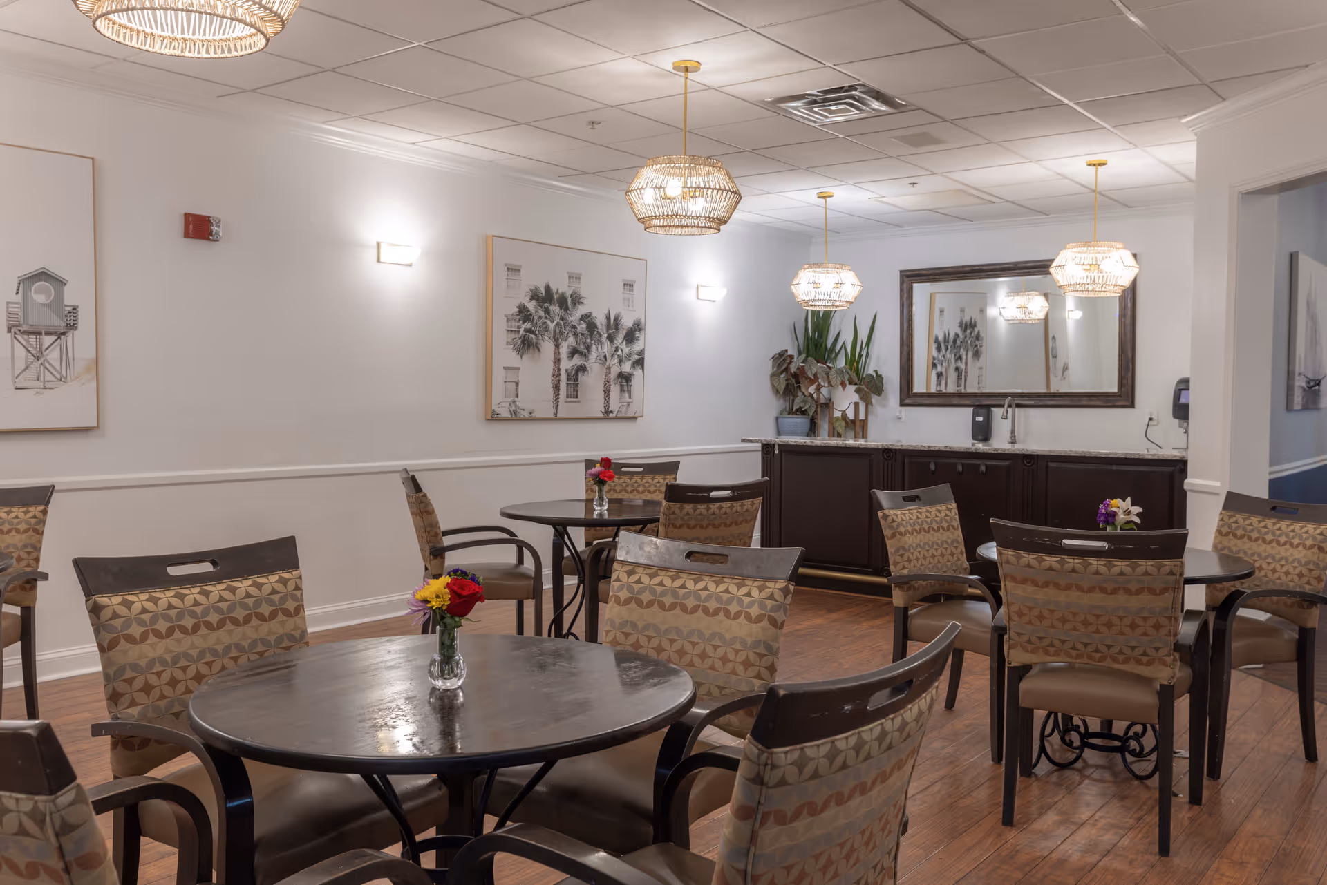 Well-lit communal dining area with round tables, patterned chairs, pendant lights, and a serving counter with mirror.