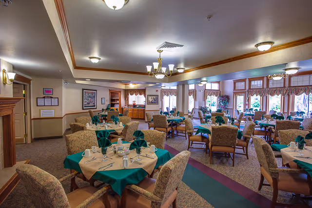 A spacious dining room with multiple round tables covered with beige tablecloths and green napkins folded elegantly. Each table is set with cups, saucers, and silverware. The room has upholstered chairs, carpeted floors, large windows with valances letting in natural light, and ceiling light fixtures including chandeliers. The walls are decorated with framed pictures and there is a wooden cabinet in the background.