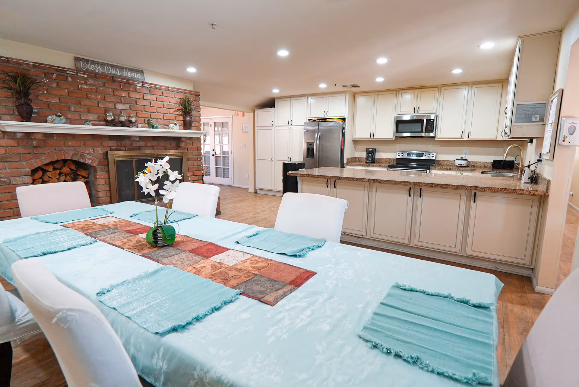 A dining area with a long table covered by a light blue tablecloth and matching placemats, with a decorative table runner and a small vase of white flowers in the center. Behind the table is a kitchen with white cabinets, a stainless steel refrigerator, microwave, stove, and granite countertops. To the left, there is a brick fireplace with a mantel decorated with plants and a sign that reads 'Bless Our Home'. The room has recessed lighting and wooden flooring.