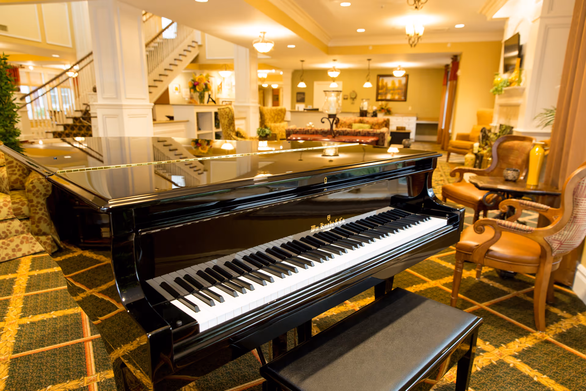 A glossy black grand piano sits in a warmly lit lobby lounge with armchairs and a staircase in the background.
