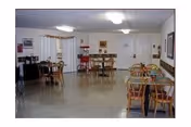 Communal dining room with several wooden tables and chairs, linoleum floor, and a serving area along the back wall.