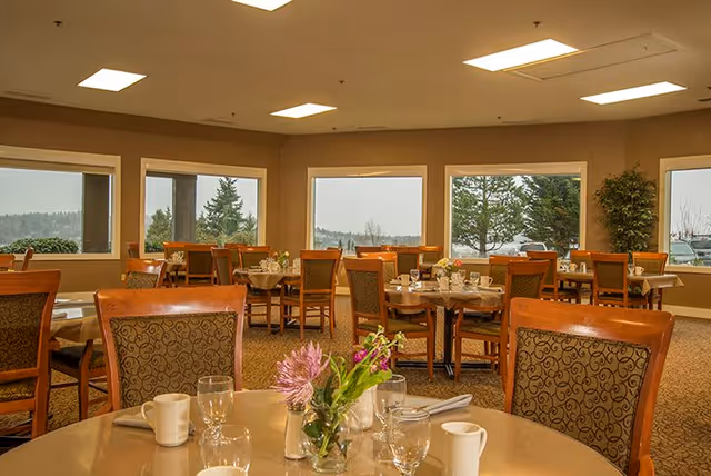 Dining room with multiple round tables set with cups, glasses, napkins, and small flower arrangements. Large windows show an outdoor view with trees and a cloudy sky. The room has warm lighting and carpeted floors.