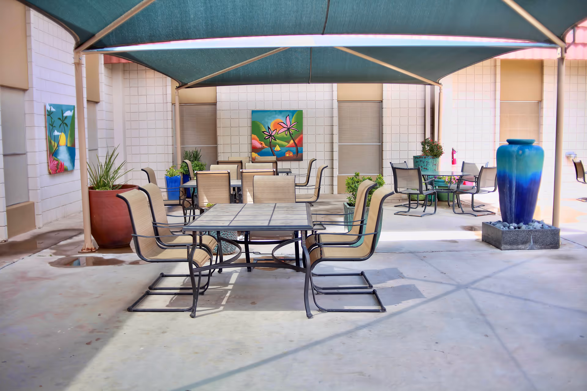 Shaded outdoor courtyard with tables and chairs, potted plants, colorful wall art, and a large blue decorative vase.