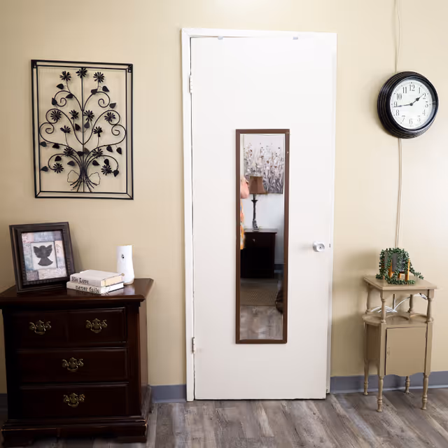 Interior view of a room with a white door featuring a narrow vertical mirror. To the left of the door is a dark wooden dresser with three drawers, holding a framed picture, two stacked books, and a small white device. Above the dresser is a decorative black metal wall art with a floral design. To the right of the door is a small beige side table with a green plant and a small framed picture. A round black wall clock showing the time as 2:50 is mounted on the wall above the side table. The floor has a wood-like finish.