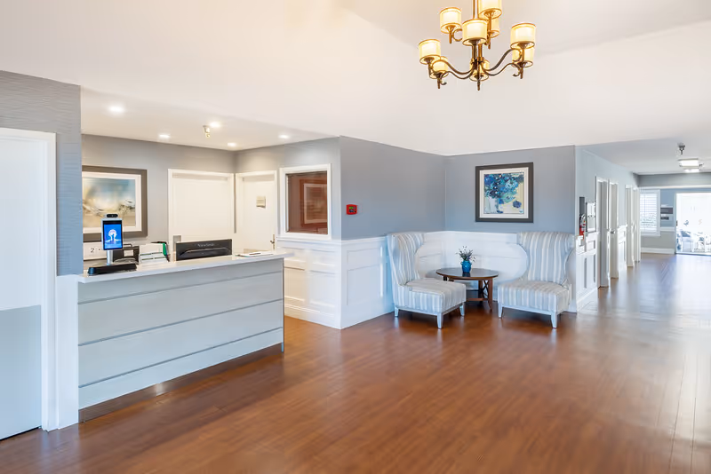 A bright and clean reception area in a senior living facility with a light gray reception desk, two striped armchairs with a small round table between them, a framed artwork on the wall, and a chandelier hanging from the ceiling. The floor is wooden, and there is a hallway leading to other rooms in the background.