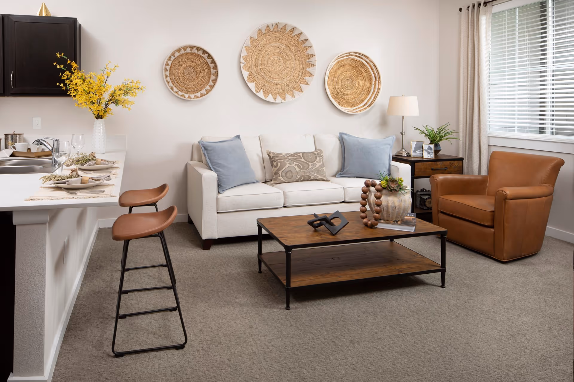 A cozy living room area with a beige sofa adorned with two blue and one patterned pillow, a brown leather armchair, and a wooden coffee table decorated with a vase and ornamental beads. The wall behind the sofa features three round woven wall hangings. To the left, there is a kitchen counter with two brown bar stools, a vase with yellow flowers, and place settings with plates and wine glasses. A window with blinds and beige curtains is on the right side, next to a side table with a lamp, plant, and framed photos.
