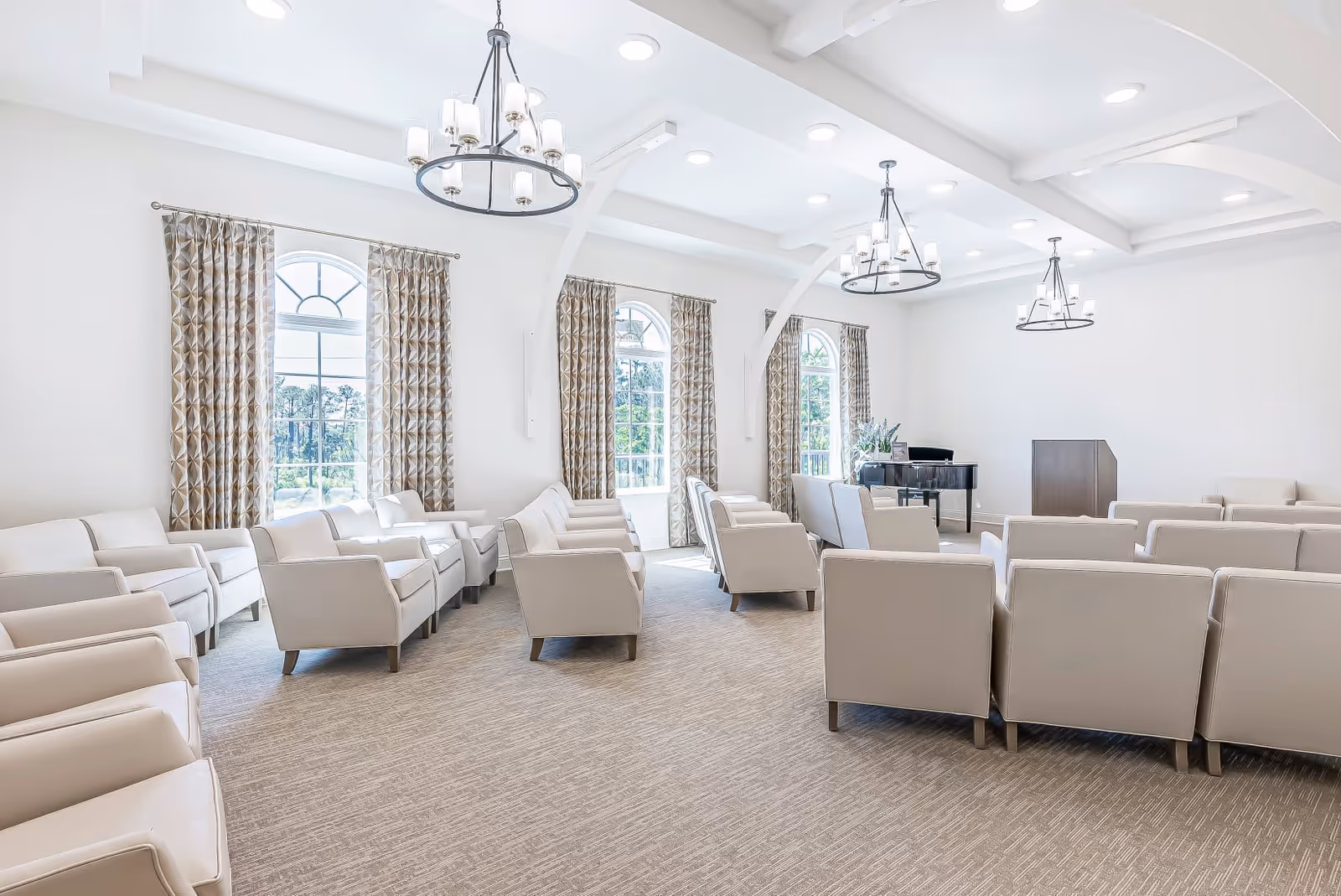 Bright communal meeting room with rows of beige armchairs, chandeliers, arched windows with patterned curtains, a piano and podium.