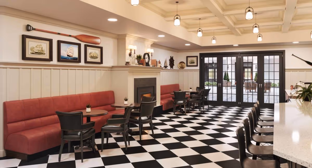 A spacious and well-lit common area with black and white checkered flooring, red cushioned bench seating along the walls, small round tables with black chairs, a fireplace with decorative items on the mantel, framed nautical artwork, and large glass doors leading to an outdoor patio.