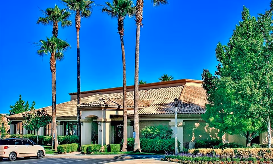 Front view of a single-story Mediterranean-style building with tall palm trees, landscaped shrubs, and a parked car.