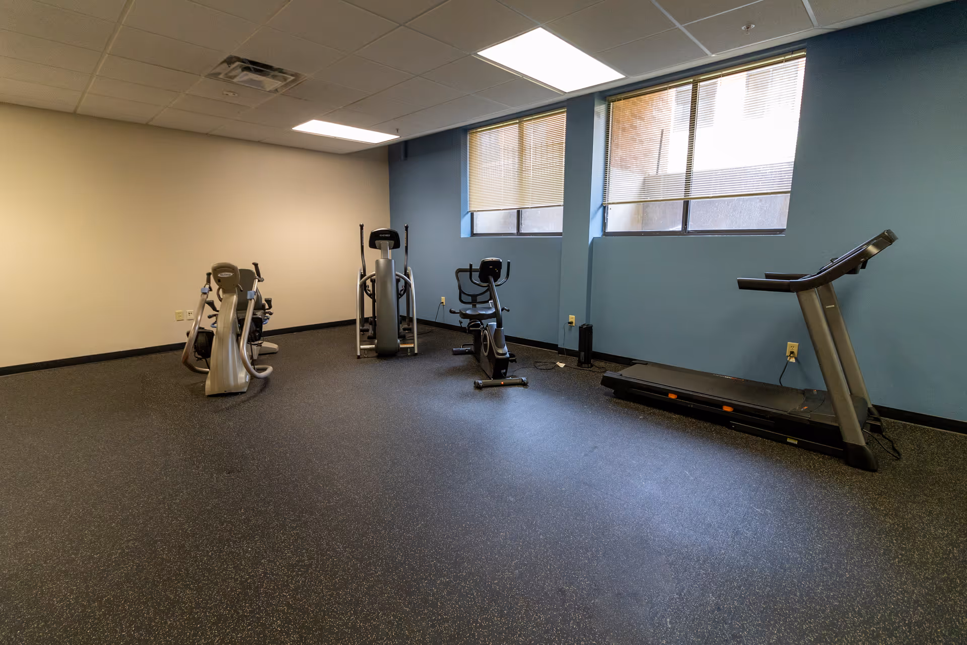 A small fitness room with exercise equipment including two stationary bikes, an elliptical machine, and a treadmill. The room has blue and beige walls, two windows with blinds partially closed, and a speckled rubber floor.