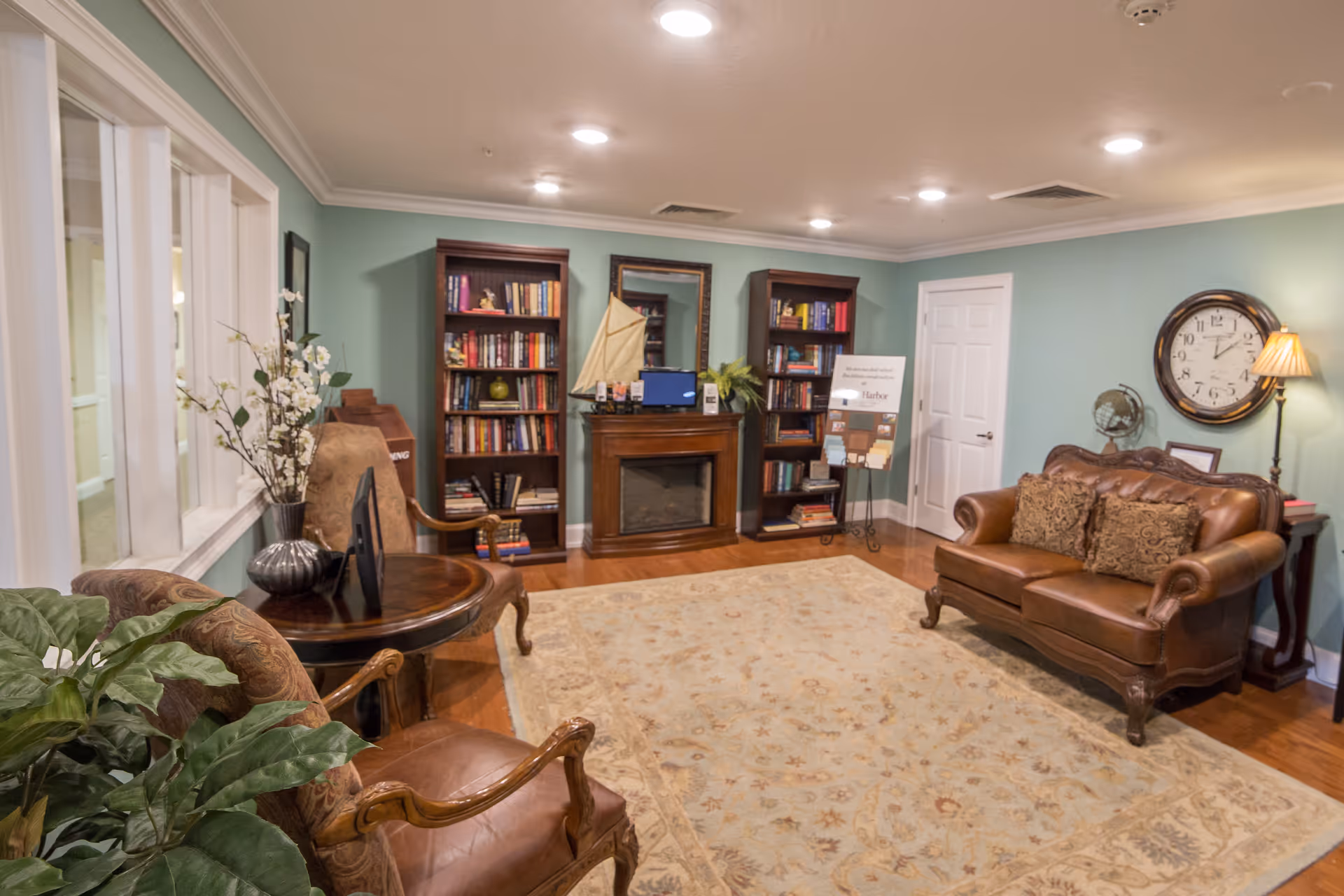 A cozy living room with light blue walls, a large beige patterned rug, a brown leather sofa with decorative pillows, two wooden bookshelves filled with books, a fireplace with a mirror and a model sailboat on the mantel, a round wooden table with a vase of flowers, and a large wall clock above a side table with a lamp and globe.