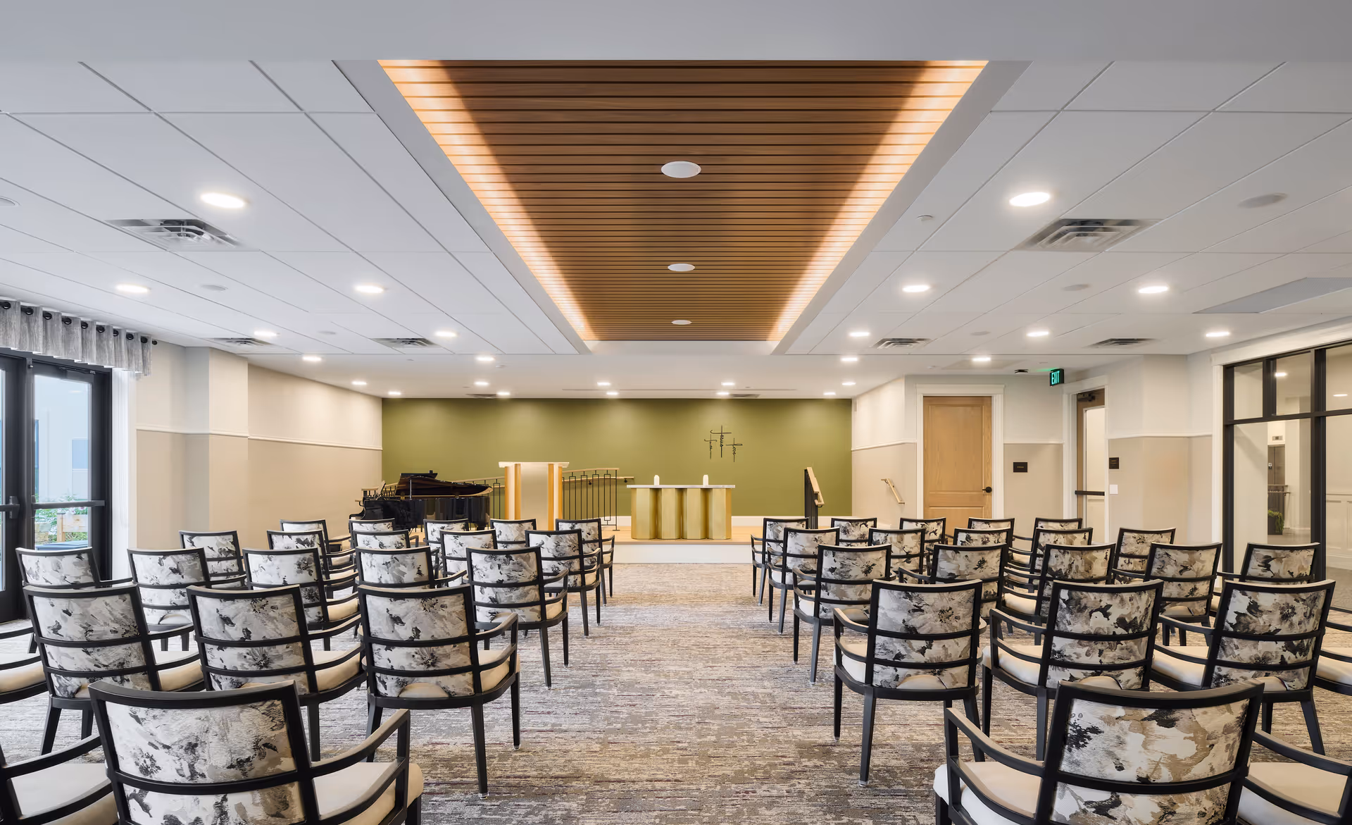 A modern, well-lit meeting or chapel room with rows of patterned chairs facing a small stage area with a podium, altar, and a piano on the left side. The ceiling features recessed lighting and a wooden panel accent in the center. There are large windows on the left side allowing natural light to enter.