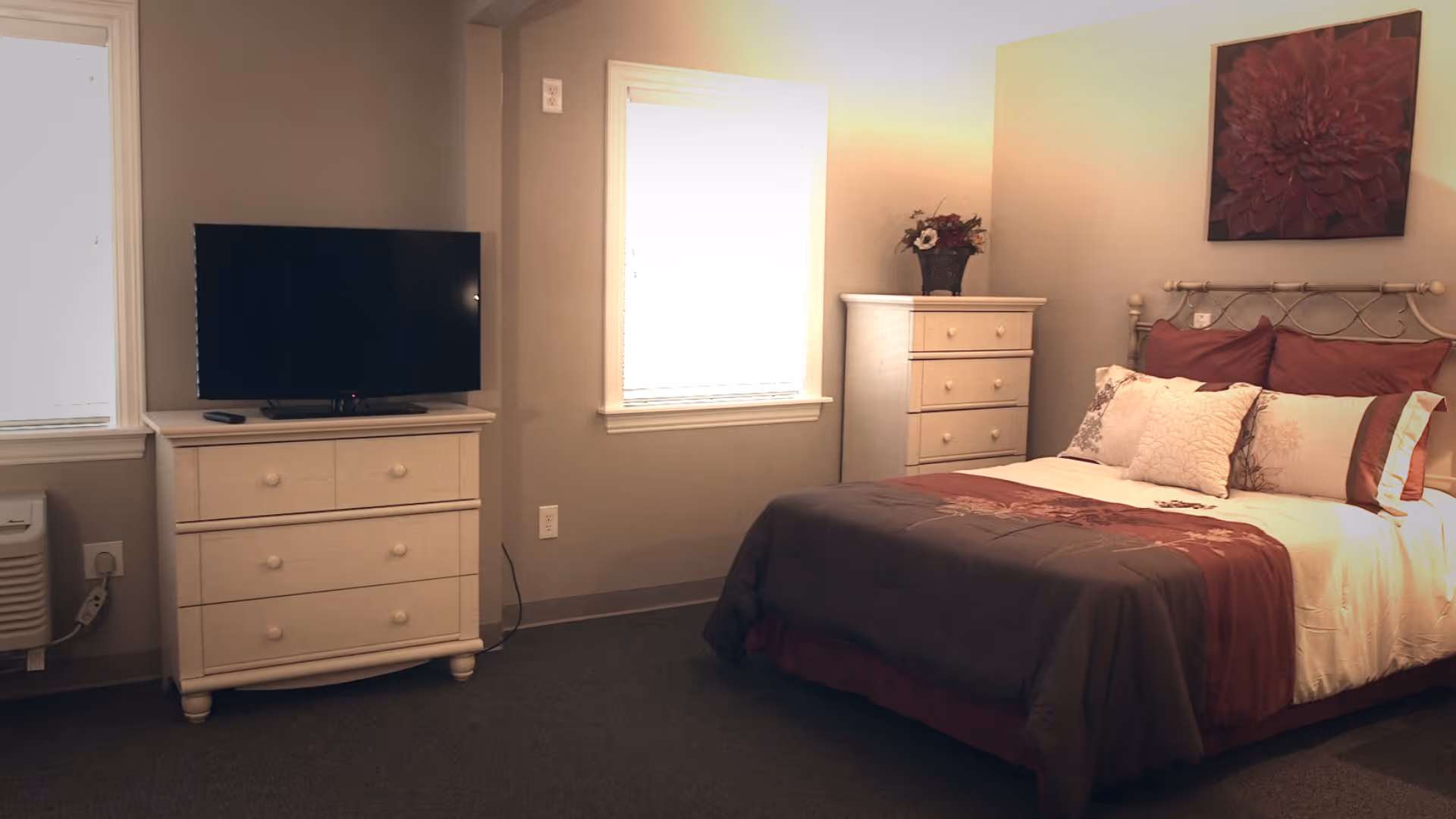 A tidy bedroom with a made bed, two white dressers, and a television on a dresser between two windows.