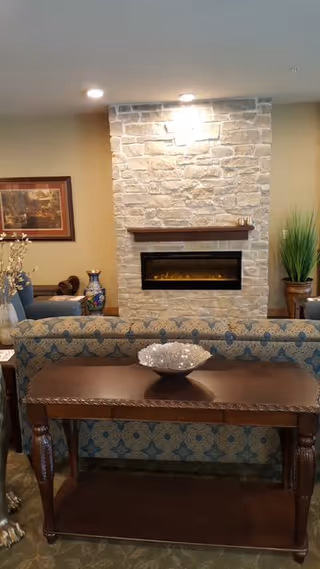 A cozy living room area featuring a stone fireplace with a wooden mantel, a patterned sofa facing away from the camera, a wooden console table with a decorative bowl on top, a framed painting on the wall, a large decorative vase, and a tall potted plant.