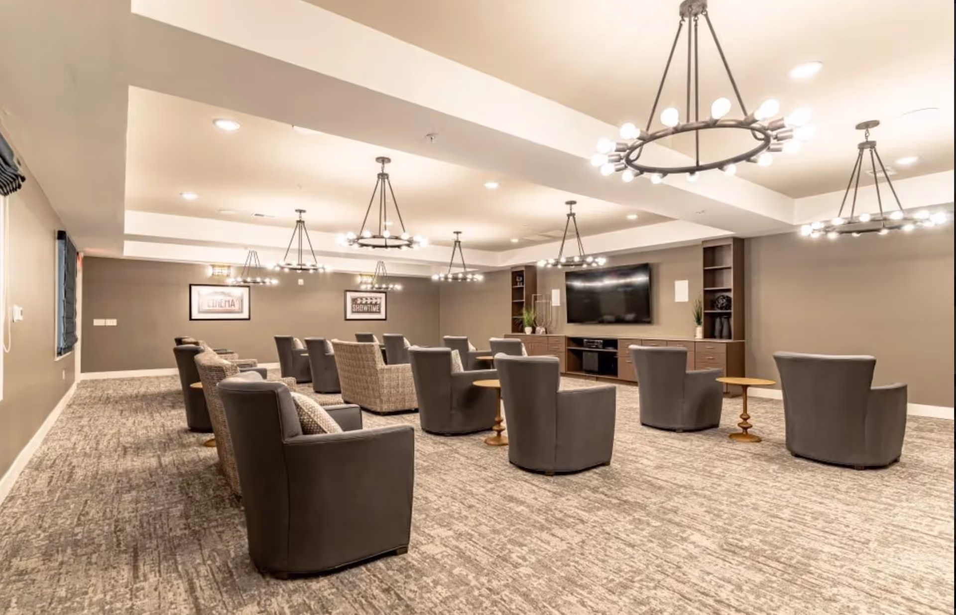 A spacious senior living common area with multiple comfortable armchairs arranged in rows facing a large flat-screen TV mounted on the wall. The room features modern circular chandeliers, neutral-colored walls, carpeted flooring, and built-in shelving units on either side of the TV.