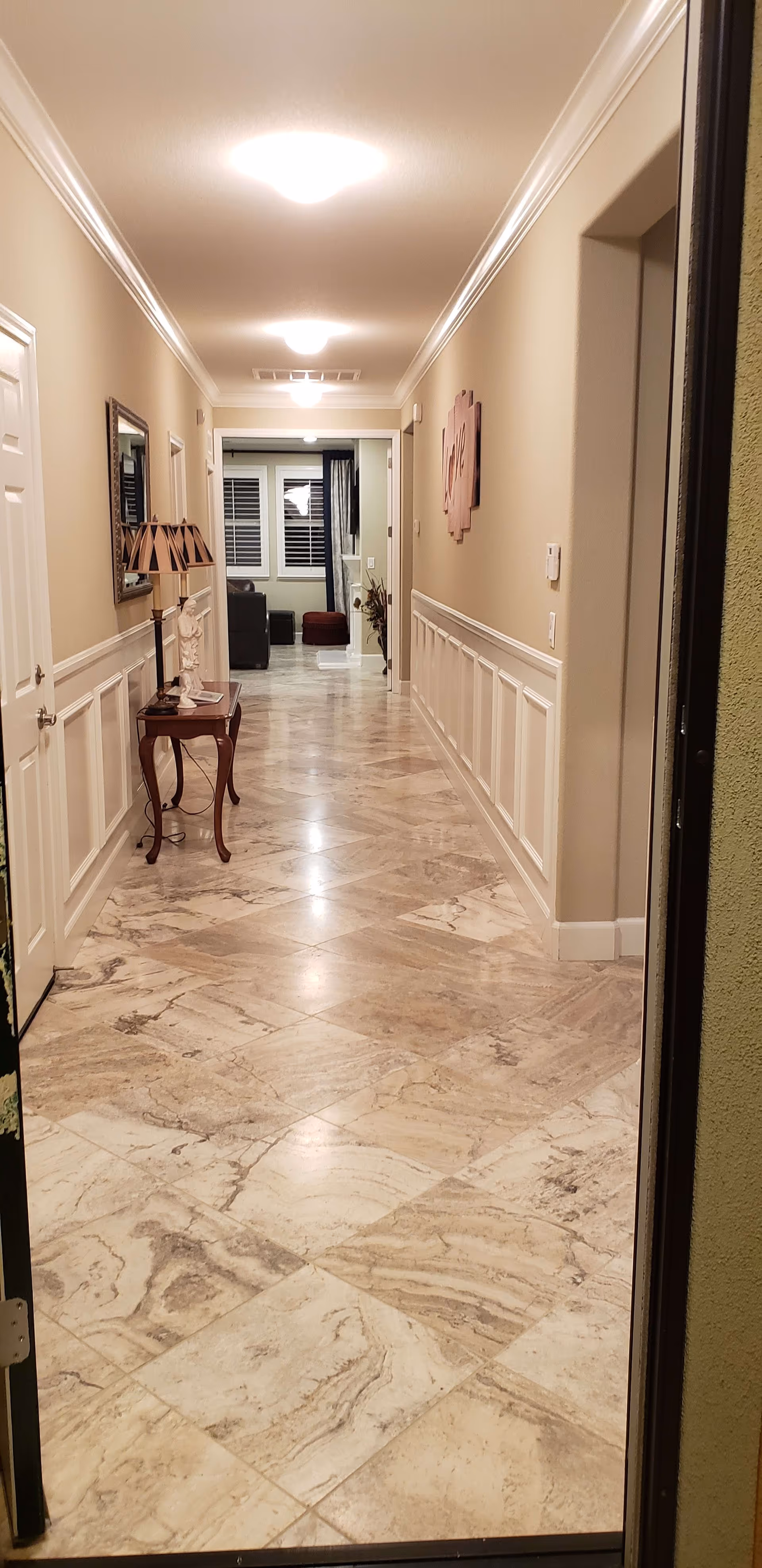 View down a long, well-lit hallway with beige walls and marble tile flooring. On the left side, there is a small wooden table with a lamp and a decorative statue, and a mirror hanging above it. At the end of the hallway, a living room area is visible with a window and furniture. A decorative wall piece with the word 'Love' is mounted on the right wall.