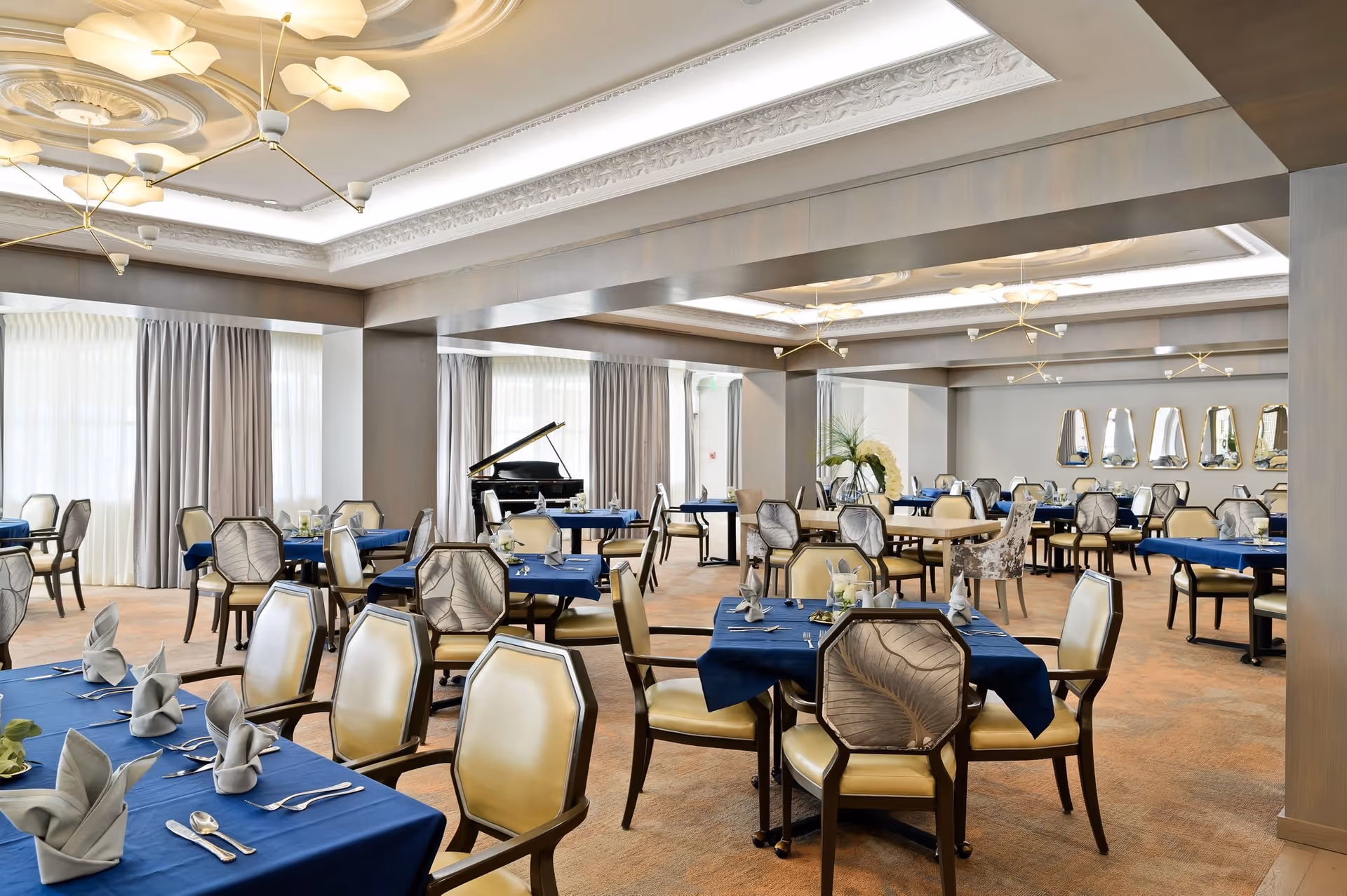A spacious and elegant dining room with multiple tables covered in blue tablecloths, each set with silverware and napkins folded in a decorative manner. The room features beige upholstered chairs with wooden frames, large windows with light curtains, modern ceiling lights, and a grand piano in the background. Mirrors and decorative plants add to the sophisticated ambiance.