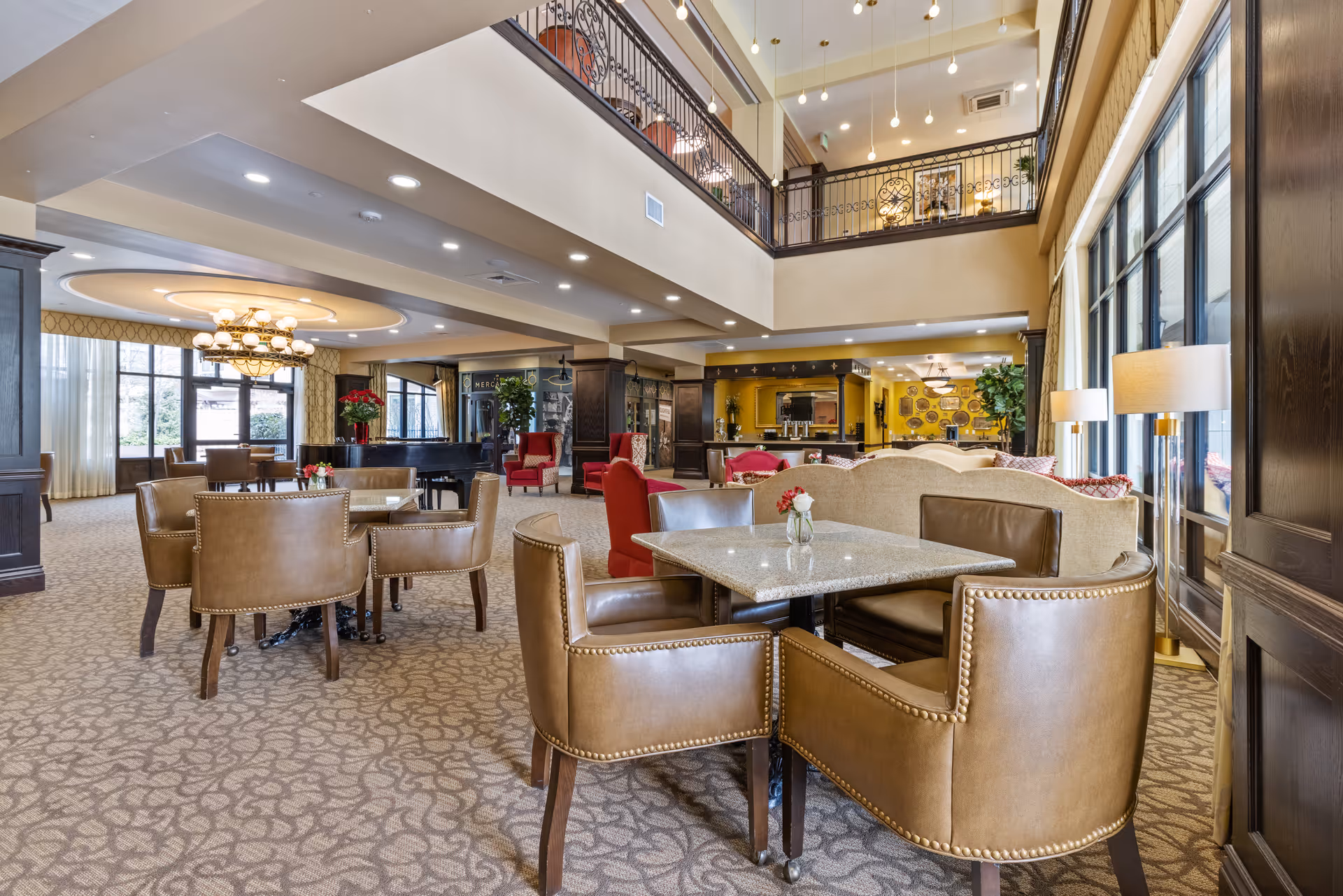 Open two-story common room with dining tables, leather chairs, sofas, and a mezzanine overlooking large windows.