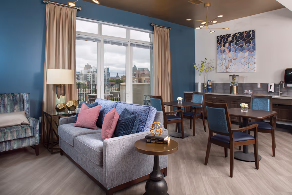 A cozy living room area with a gray sofa adorned with pink and blue pillows, a patterned armchair, and a small round side table with decorative items. Behind the sofa, there are two small dining tables with blue chairs. The room features large windows with beige curtains, offering a view of a cityscape outside. The walls are painted blue and white, with a geometric artwork hanging on the white wall. A countertop with a water dispenser and glasses is visible in the background.