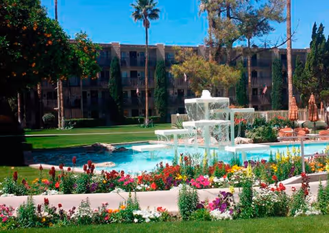 Outdoor garden area with a multi-tiered white fountain surrounded by colorful flowers and green grass. In the background, there is a multi-story building with balconies and tall trees under a clear blue sky.