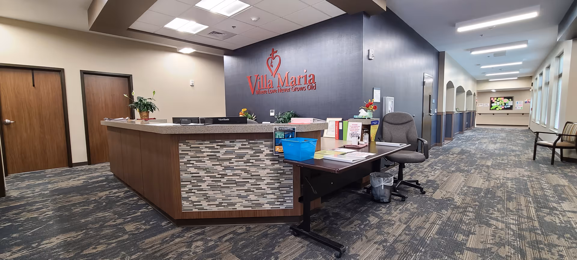Reception area of Villa Maria facility with a front desk, office chair, and a sign on the wall that reads 'Villa Maria Where Love Never Grows Old'. The area has carpeted flooring, two closed wooden doors on the left, and a long hallway with windows and chairs on the right.