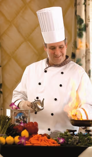A chef wearing a white double-breasted chef coat and tall white chef hat is cooking with a pan that has flames rising from it. The chef is smiling and holding a small metal container, possibly pouring oil or another ingredient into the pan. In front of the chef, there is a table with various fresh vegetables including carrots, lemons, and red bell peppers, along with some purple flowers and greenery. The background features a patterned wall and a curtain with a floral design.