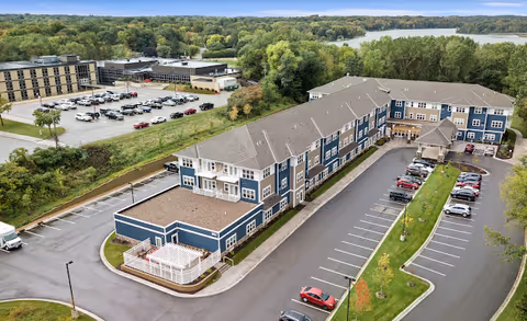 Aerial view of a multi-story blue senior living building with parking lots, driveways, and trees with a lake in the background.