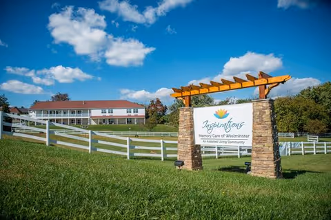 A large green lawn with a white fence leading up to a two-story building under a blue sky with scattered clouds. In the foreground, there is a stone and wood sign that reads 'Inspirations Memory Care of Westminster An Assisted Living Community.'
